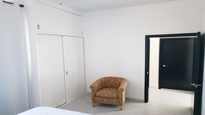 Um quarto com uma cama, um guarda-roupa, uma poltrona estampada e uma porta.