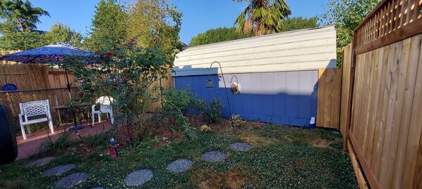 Un patio trasero con cerca de madera, un cobertizo azul y un área de patio con mesa y sillas.