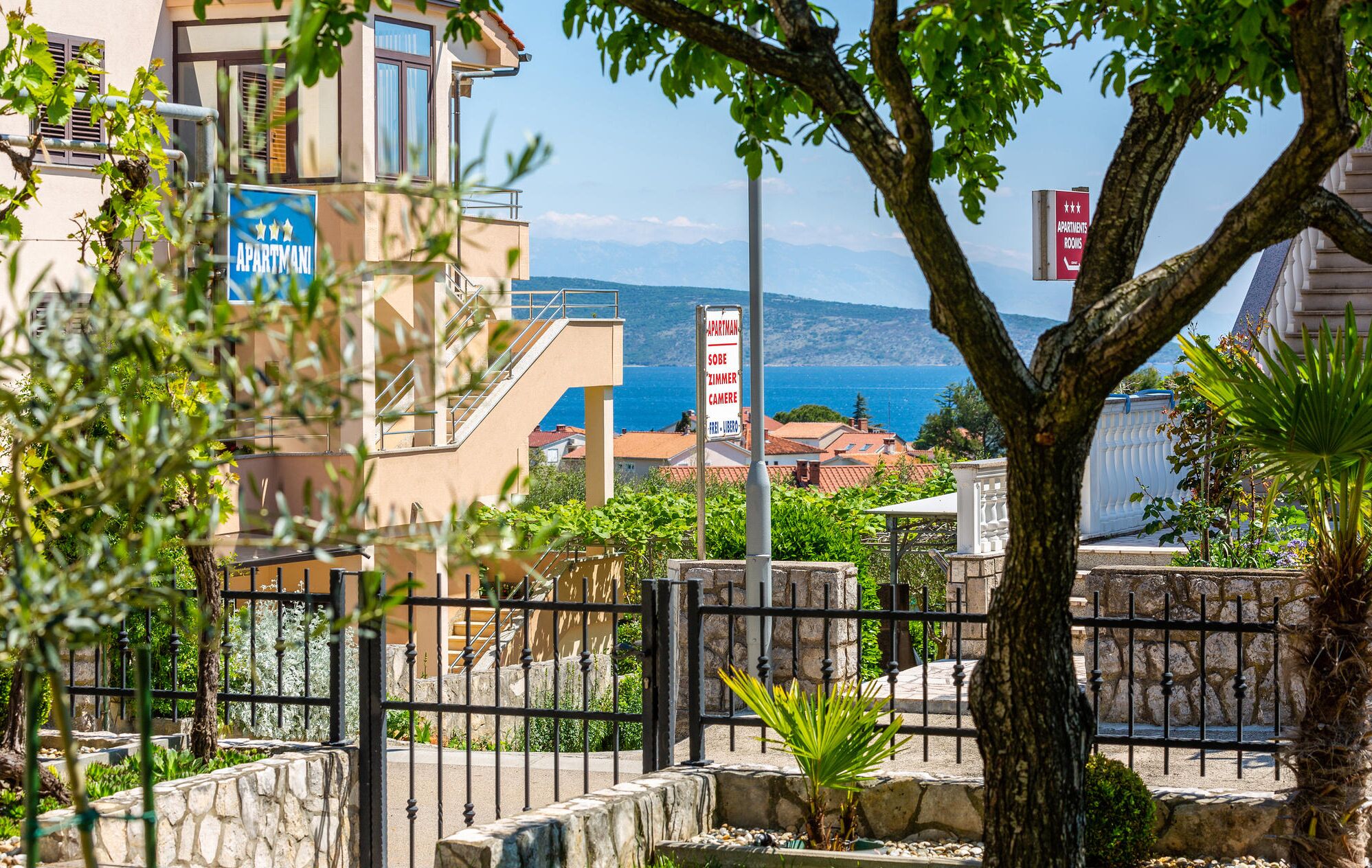 Un immeuble d’appartements situé sur la côte, avec vue sur la mer, une enseigne indiquant « Apartman » et « Sobe Zimmer Camera », et un mur de pierre orné de verdure.