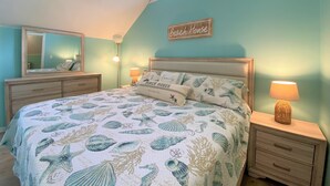 Une chambre à coucher avec un lit, des tables de chevet, une commode, un miroir et un panneau décoratif représentant une maison de plage accroché au mur.