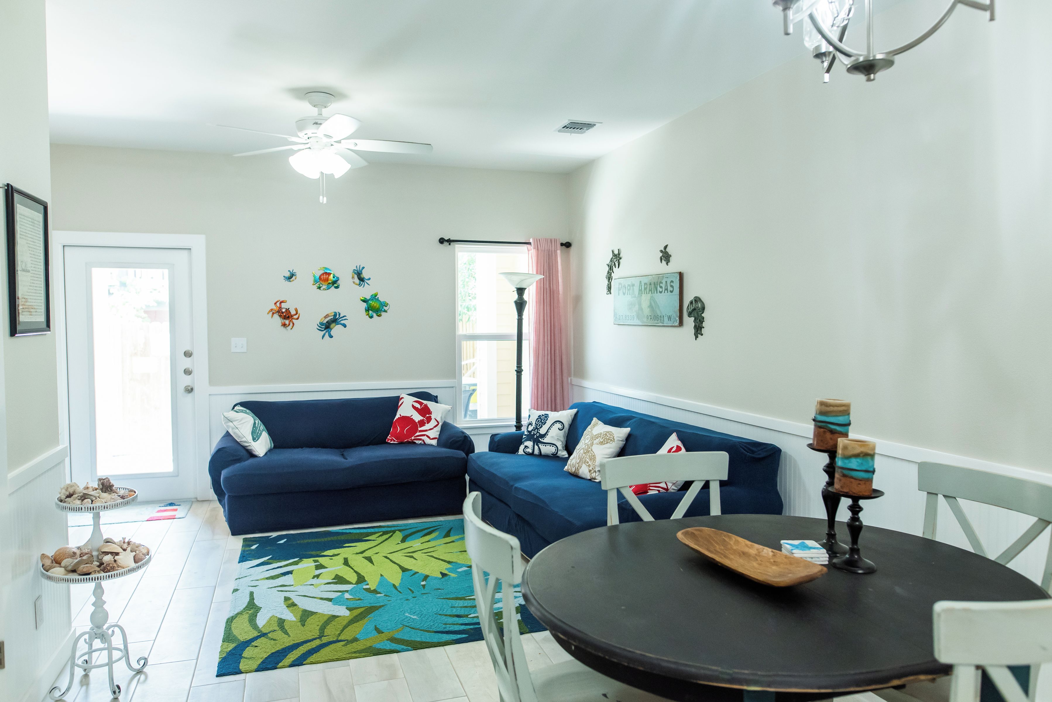 A living room with a blue sofa, a dining table with two chairs, a ceiling fan, and a colorful rug.