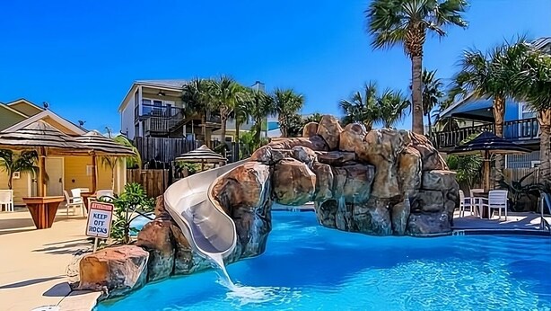Una piscina amb tobogan, palmeres i un cartell que avisa de no caminar sobre les roques.