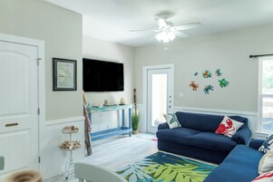Una sala de estar con un sofá azul, un televisor de pantalla plana montado en la pared, un ventilador de techo y un adhesivo decorativo en la pared con motivos marinos.