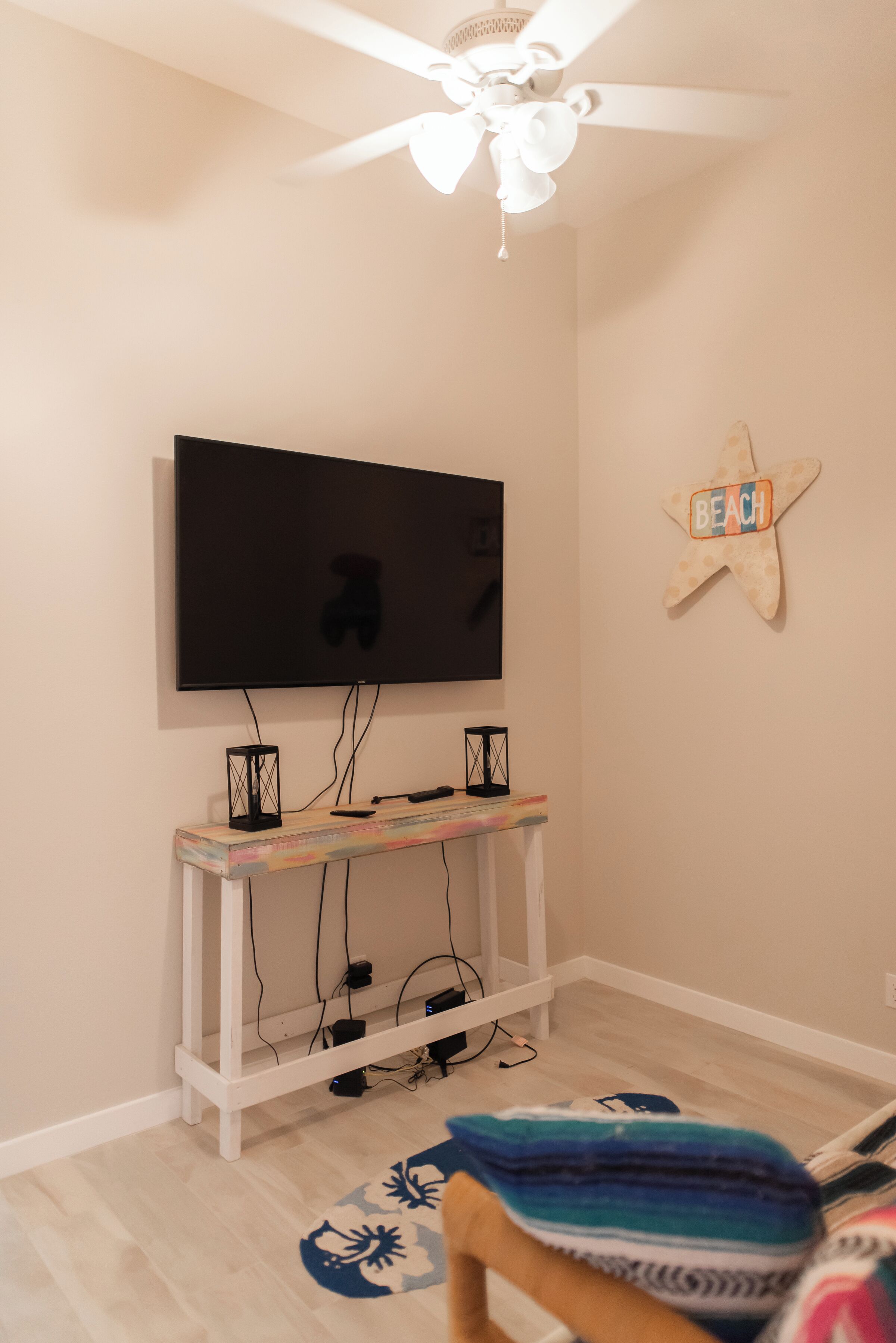 Une chambre dotée d’un téléviseur à écran plat fixé au mur, d’un ventilateur de plafond, d’une décoration en forme d’étoile de mer et d’une petite table recouverte d’une couverture colorée.