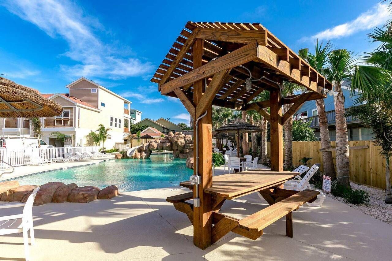 A wooden gazebo with a table and benches by a pool, surrounded by palm trees and a clear blue sky.