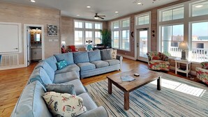A spacious living room with a large L-shaped sectional sofa, a coffee table, and a view of the city through large windows. - Turtle Paradise by Silver Sands Vacation Rentals (Port Aransas)