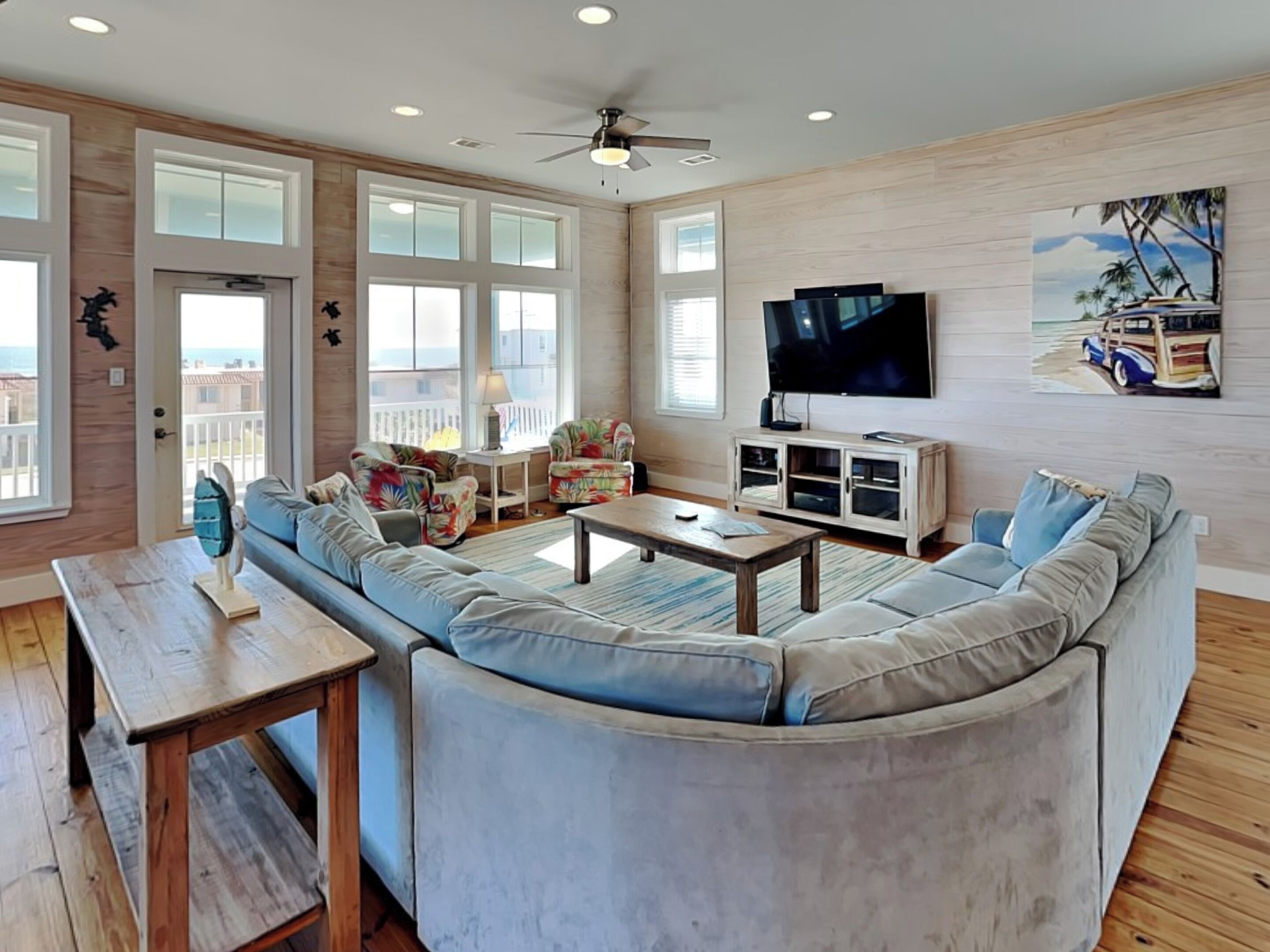A spacious living room with a large sectional sofa, a coffee table, and a TV. There are multiple windows providing natural light and a view of the outside.