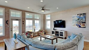 A spacious living room with a large sectional sofa, a coffee table, and a TV. There are multiple windows providing natural light and a view of the outside. - Turtle Paradise by Silver Sands Vacation Rentals (Port Aransas)