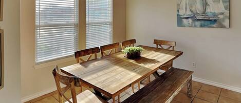 A dining room with a wooden table, chairs, a bench, and a chandelier. There are two windows with blinds, a painting of sailboats on the wall, and a potted plant on the table.