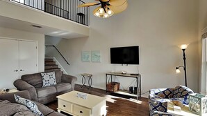 A spacious living room with a high ceiling fan, a flat-screen TV mounted on the wall, and a staircase leading to an upper level. - Sol Searcher by Silver Sands Vacation Rentals (Port Aransas)