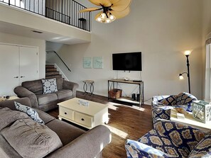 Un vaste salon doté d’un haut ventilateur de plafond, d’un téléviseur à écran plat fixé au mur et d’un escalier menant à l’étage supérieur.
