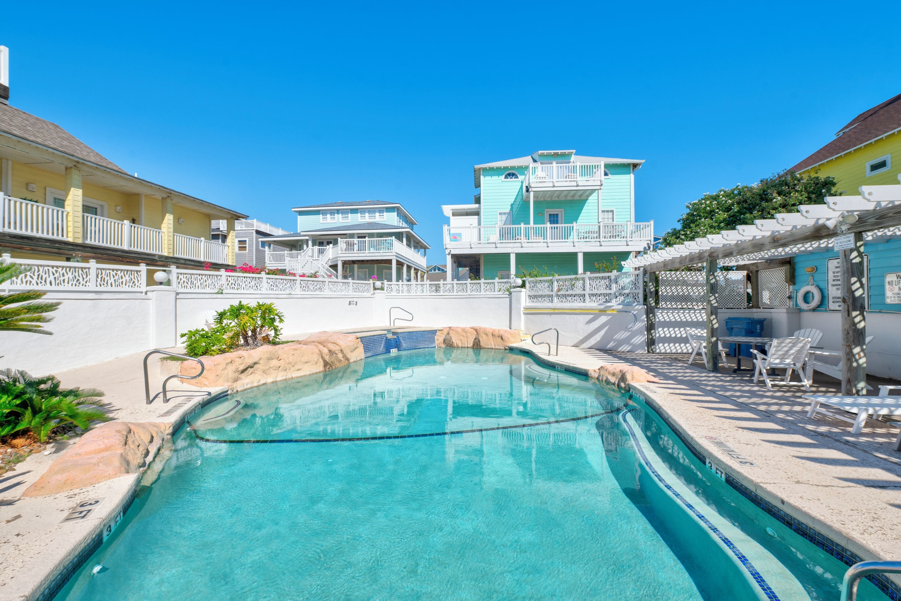 Una piscina con acqua cristallina e blu, circondata da una recinzione bianca e un edificio con numerosi balconi.