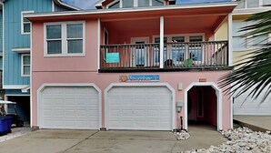 A two-story house with a pink facade, two white garage doors, and a balcony with a metal railing. - Sol Searcher by Silver Sands Vacation Rentals (Port Aransas)