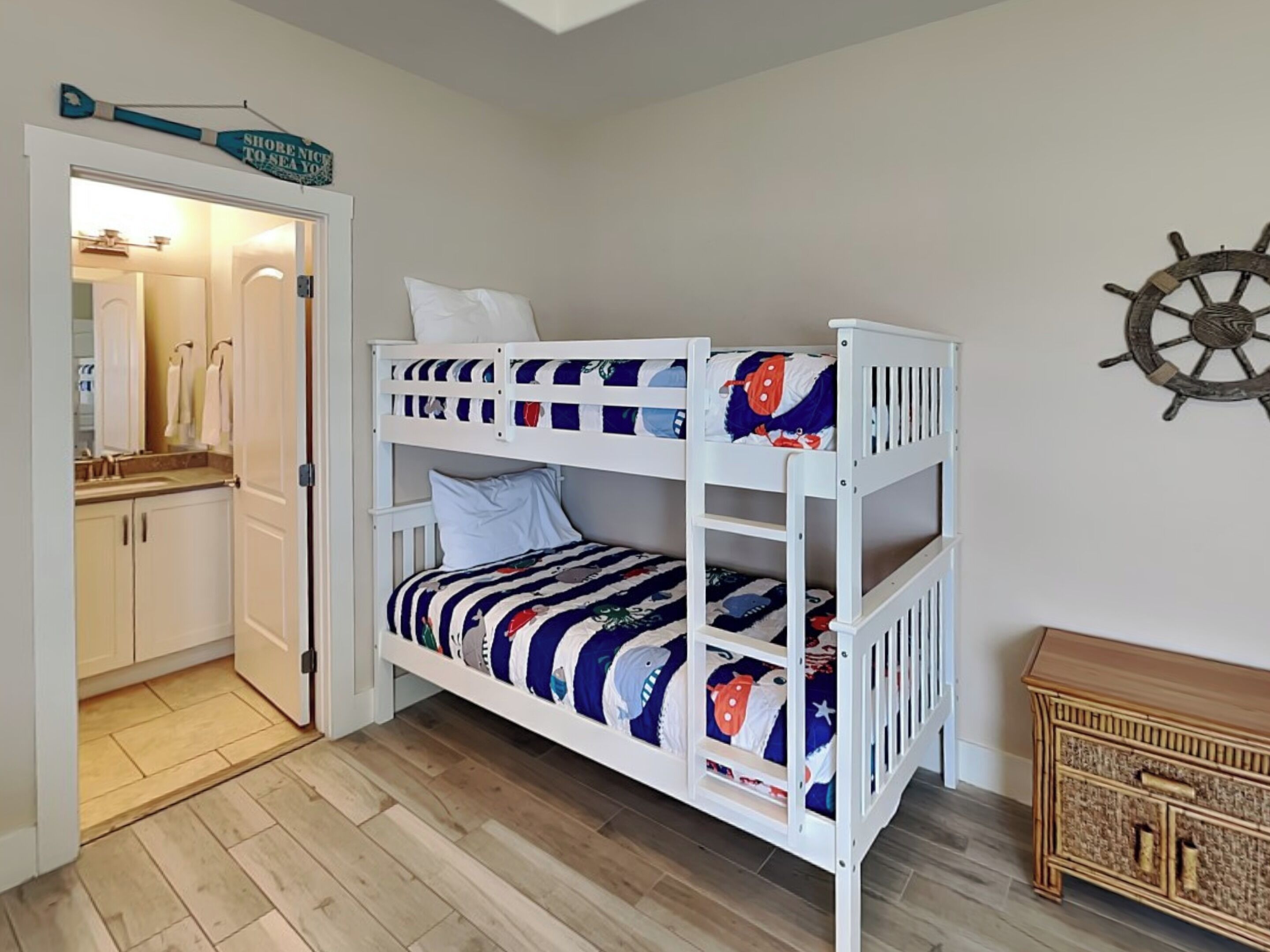 Un lit superposé avec une couvre-lit sur le thème de la marine, une commode en bois et une décoration représentant un gouvernail de navire accrochée au mur.