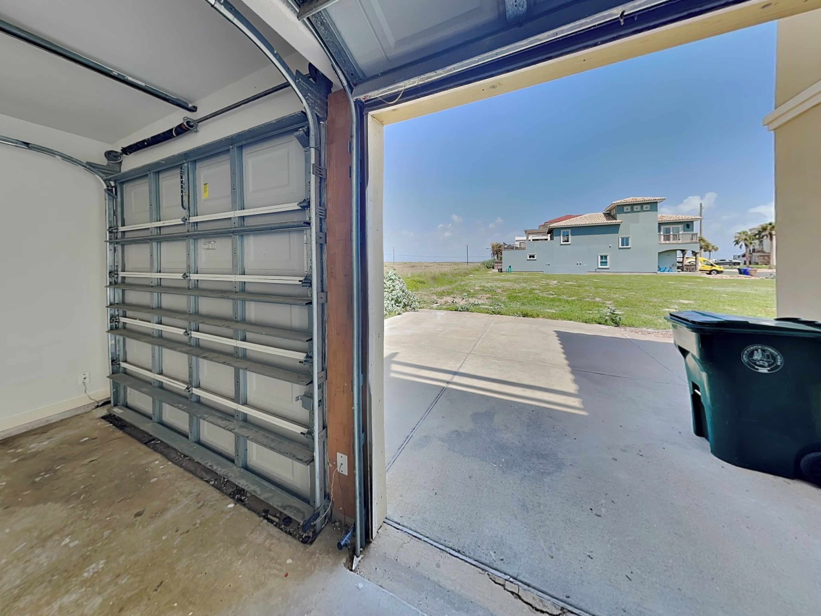 A garage with a metal door, a concrete floor, and a green trash bin.