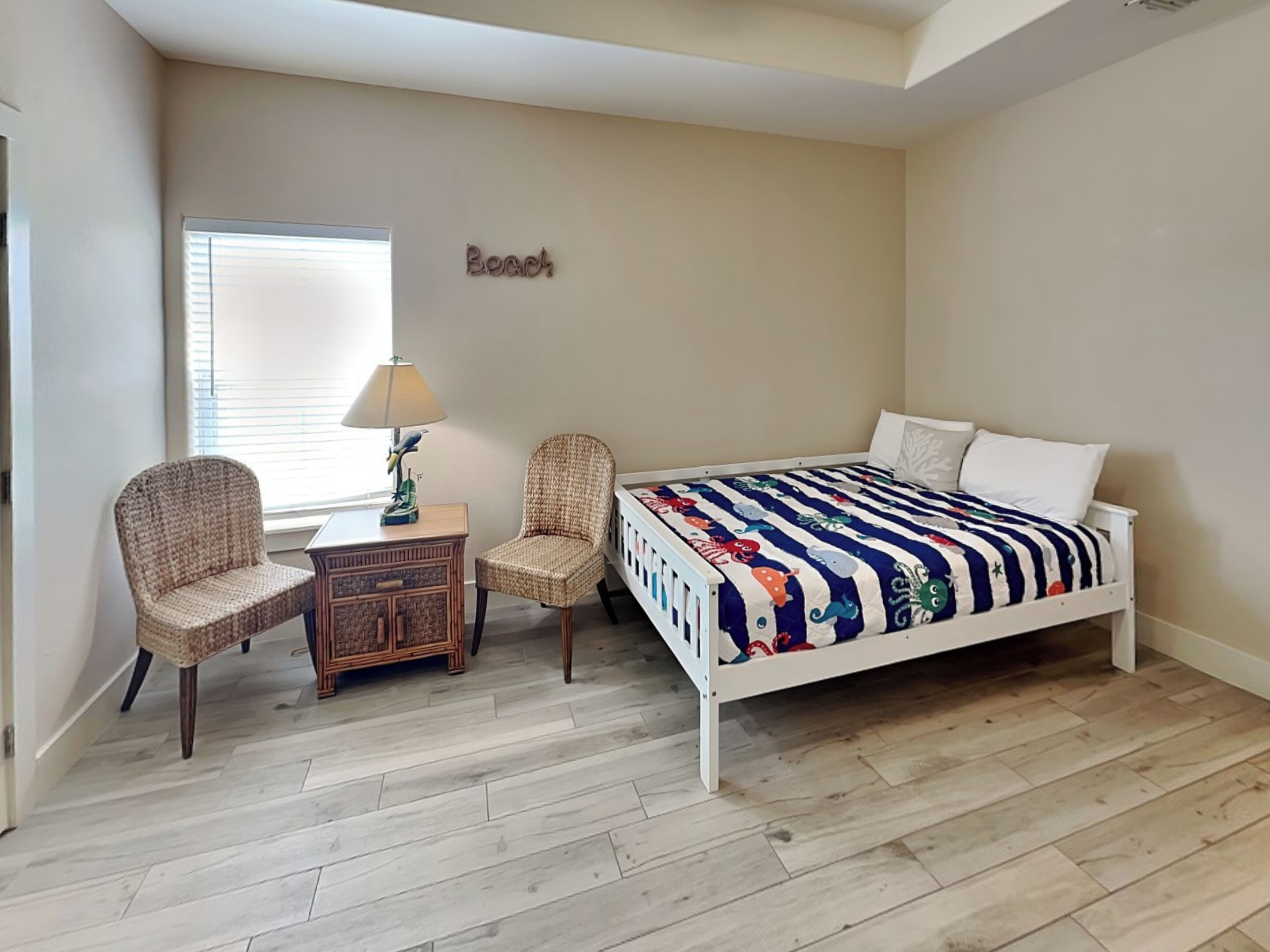 A bedroom with a bed, two wicker chairs, a small table with a lamp, and a window with blinds.