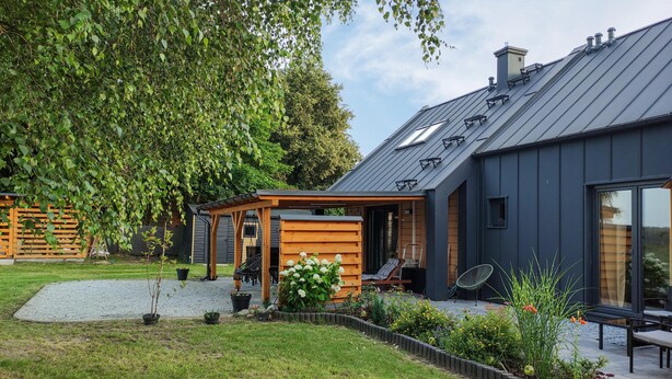 A modern house with a covered outdoor seating area, a garden with various plants, and a large tree in the background. - Cozy Home in Rudzienice (Rudzienice)