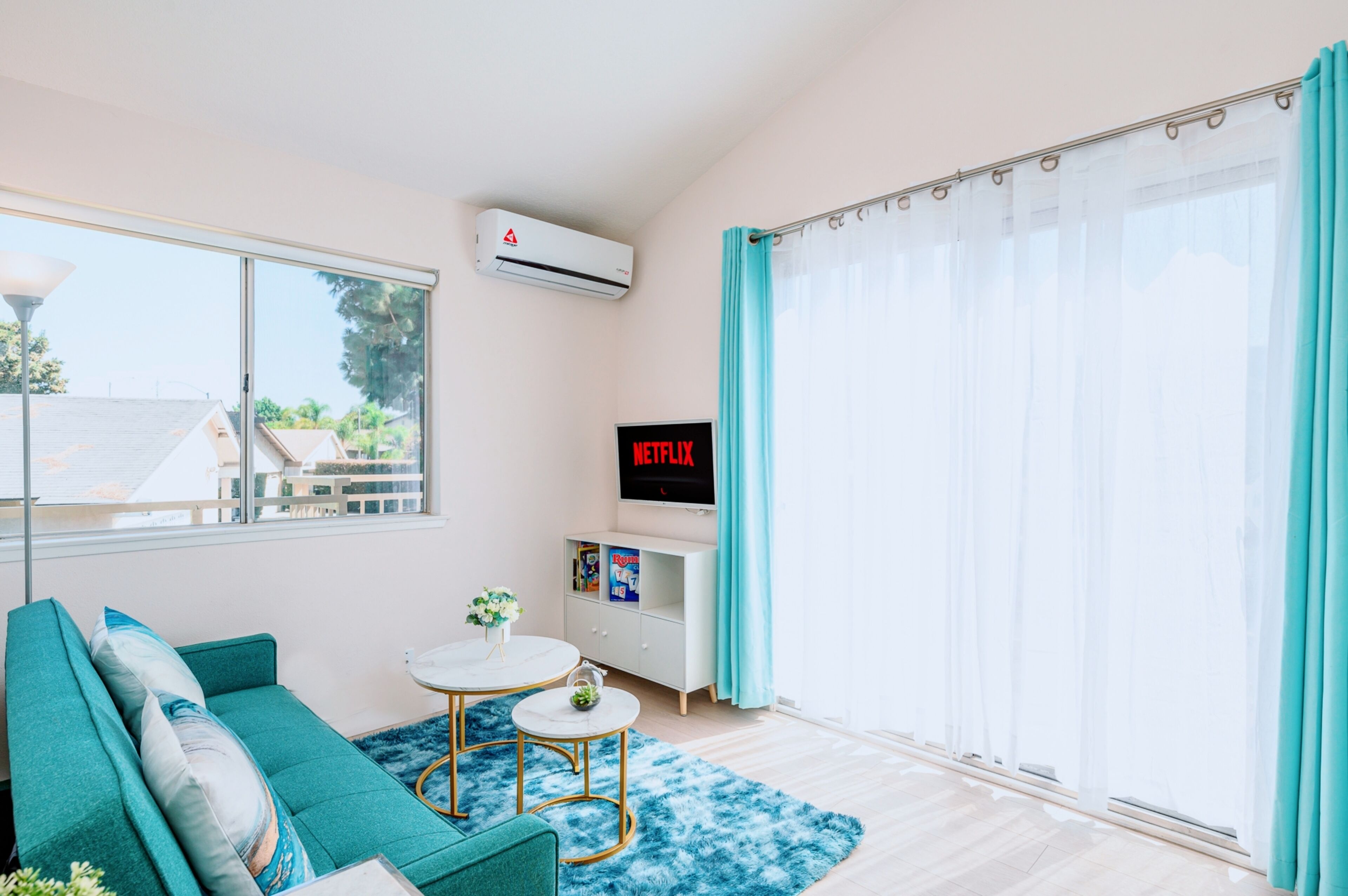 Un salon moderne avec un canapé de couleur turquoise, une petite table ronde ornée d'un vase, un meuble blanc sur lequel est posée une télévision à écran plat affichant le logo « NETFLIX », et une grande fenêtre donnant sur des immeubles et des arbres.