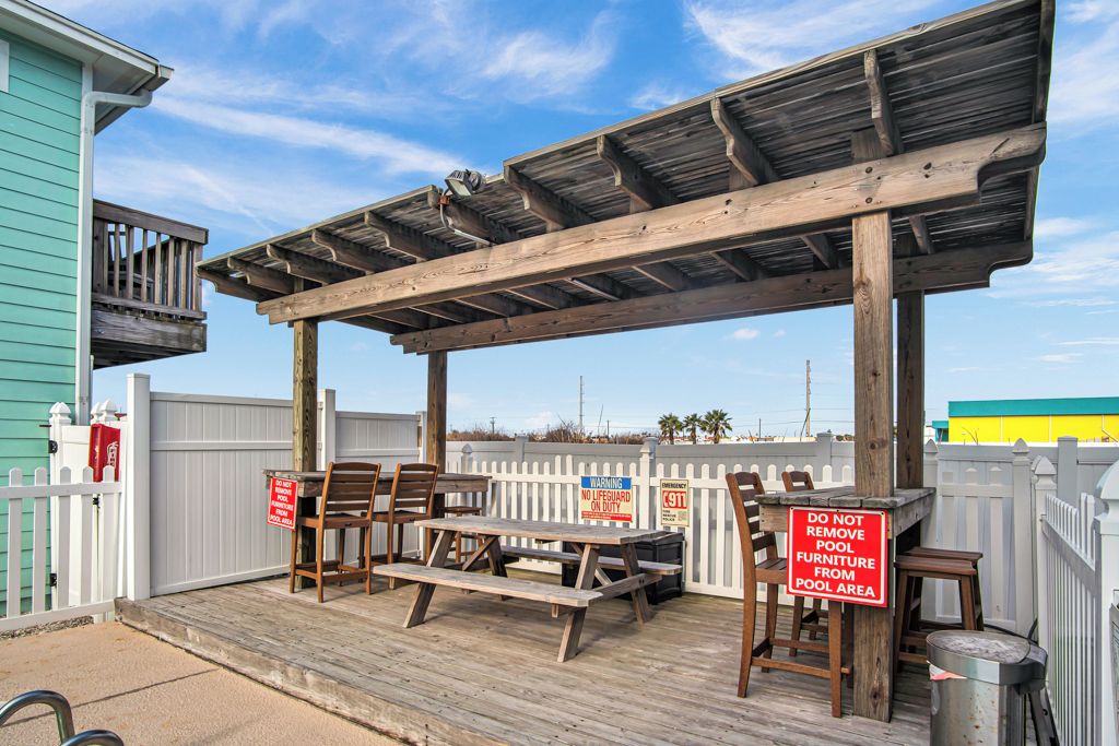 A covered outdoor seating area with picnic tables and chairs, a wooden pergola, and a white picket fence.