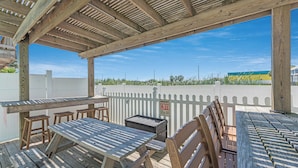 An outdoor wooden deck with picnic tables and benches, a white picket fence, and a clear blue sky. - Surfin' Seahorse by Silver Sands Vacation Rentals (Port Aransas)