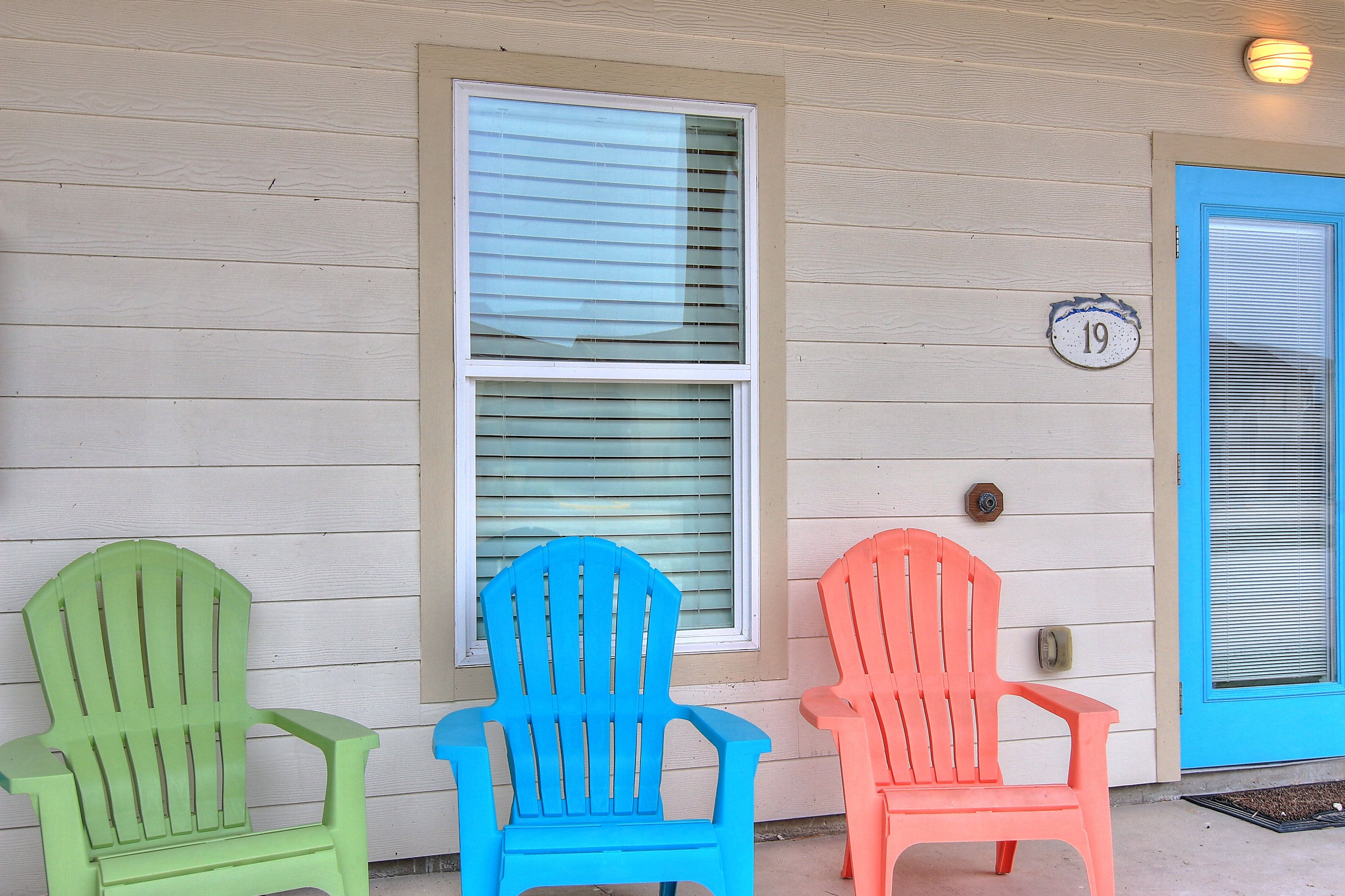 Drei Adirondack-Stühle vor einem Haus mit einer Hausnummer '19', einer blauen Tür und einem Fenster mit Rollläden.