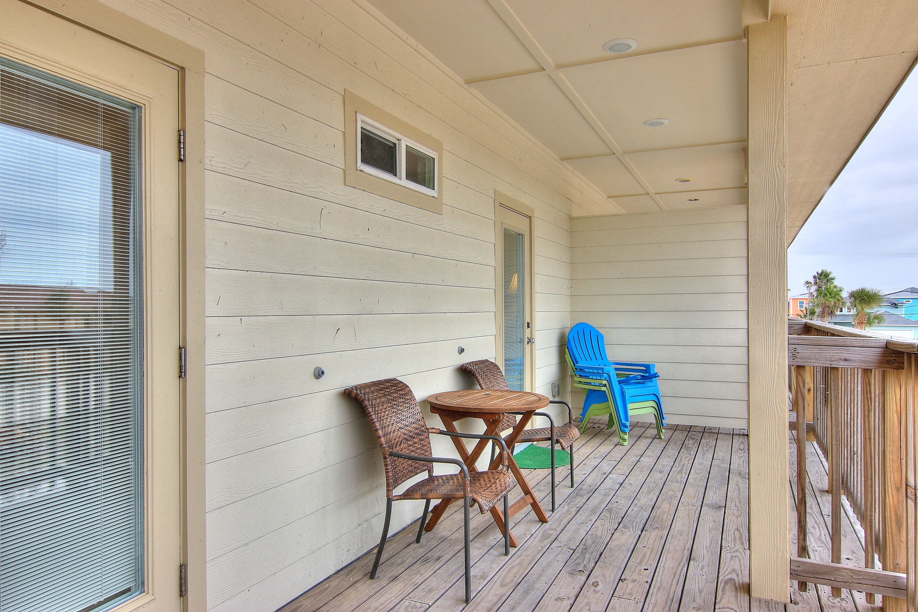 A covered porch with a wooden table and two wicker chairs, a small door, and a blue plastic chair.