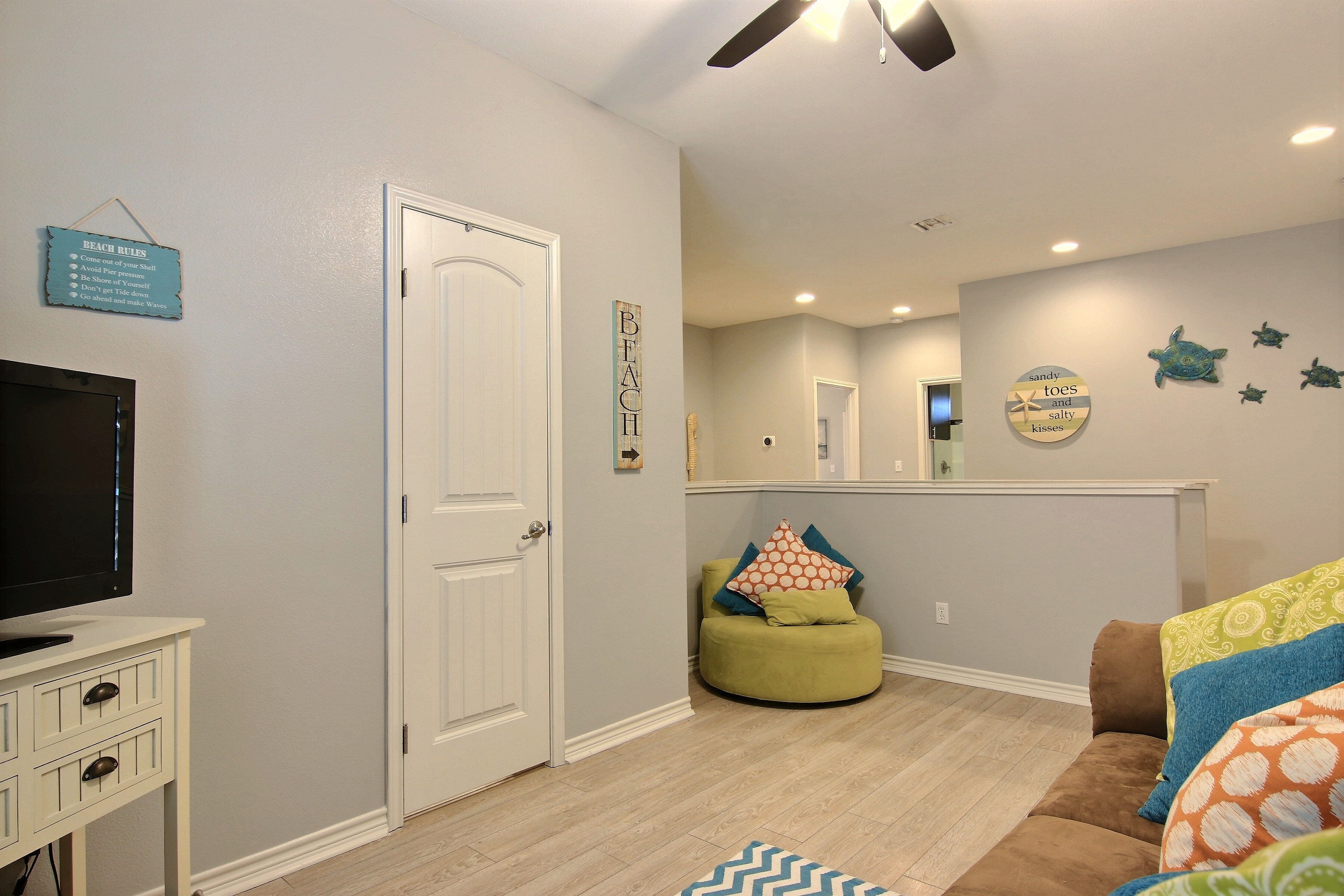 Una sala de estar con televisor, puerta, un sillón informal verde y un sofá con cojines estampados.