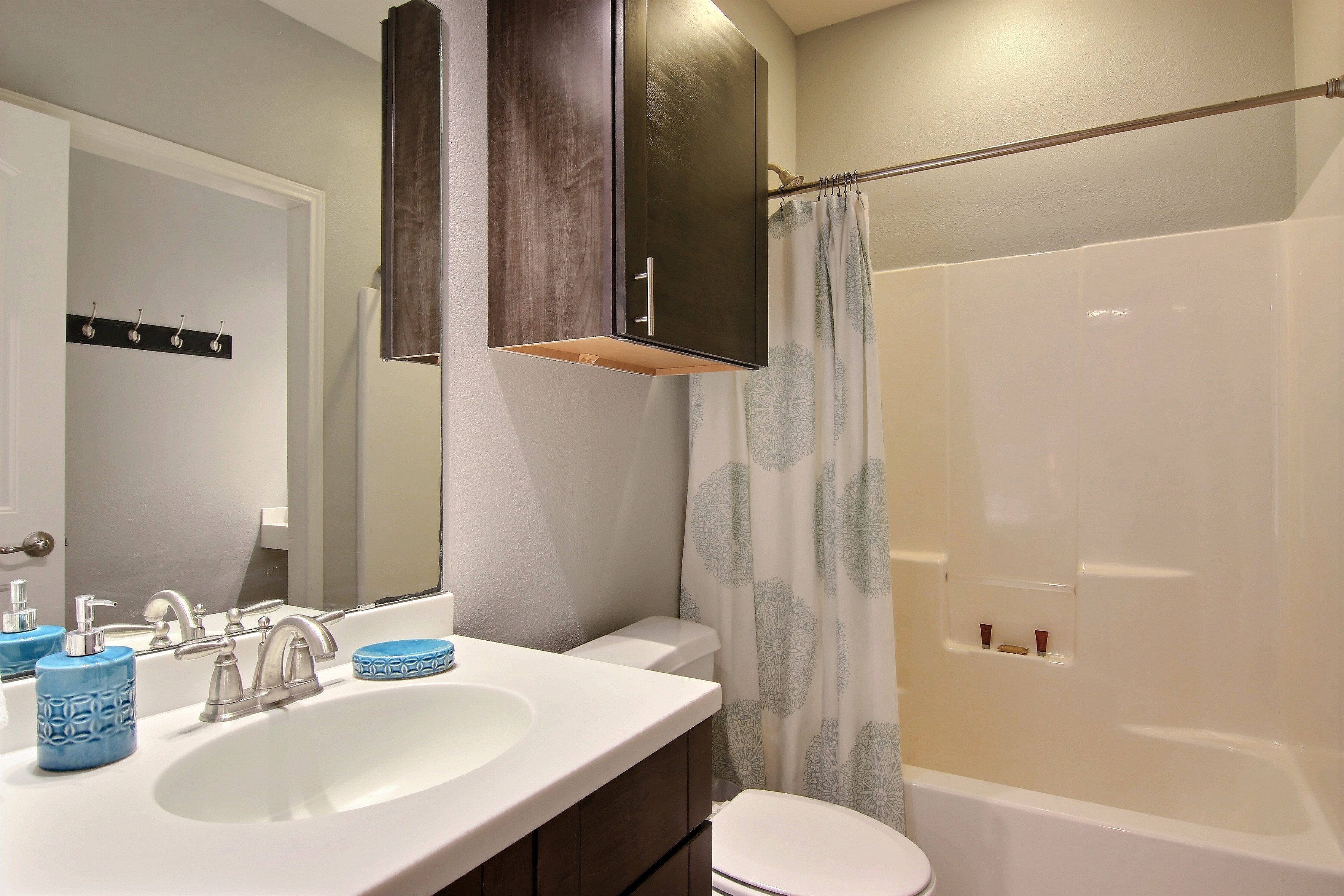 Une salle de bain avec un lavabo blanc, deux robinets, un miroir, un meuble, des toilettes et une douche avec un rideau.