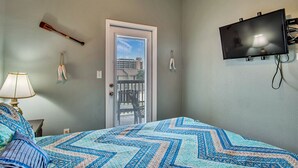 Une chambre à coucher comprenant un lit, un téléviseur fixé au mur, une porte donnant sur un balcon et une fenêtre avec des stores.