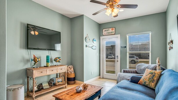 Un salon avec un canapé bleu, une table basse en bois et un ventilateur de plafond. Un téléviseur est fixé au mur, et une enseigne indique : « JUST COAST ».