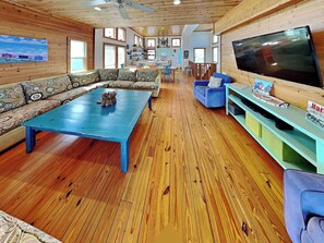 A spacious living room with a large blue coffee table, a sofa set, a television, and a kitchen area visible in the background. - The Fire Escape by Silver Sands Vacation Rentals (Port Aransas)