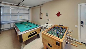 A room with a pool table and a foosball table, a garage door, and a wall-mounted fire extinguisher. - The Fire Escape by Silver Sands Vacation Rentals (Port Aransas)