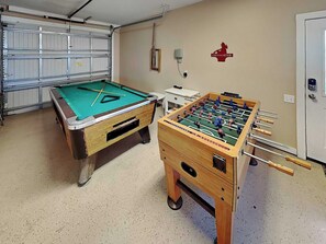 A room with a pool table and a foosball table, a garage door, and a wall-mounted fire extinguisher.