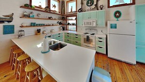 A kitchen with a central island, wooden stools, and a view of the beach through the windows. - The Fire Escape by Silver Sands Vacation Rentals (Port Aransas)