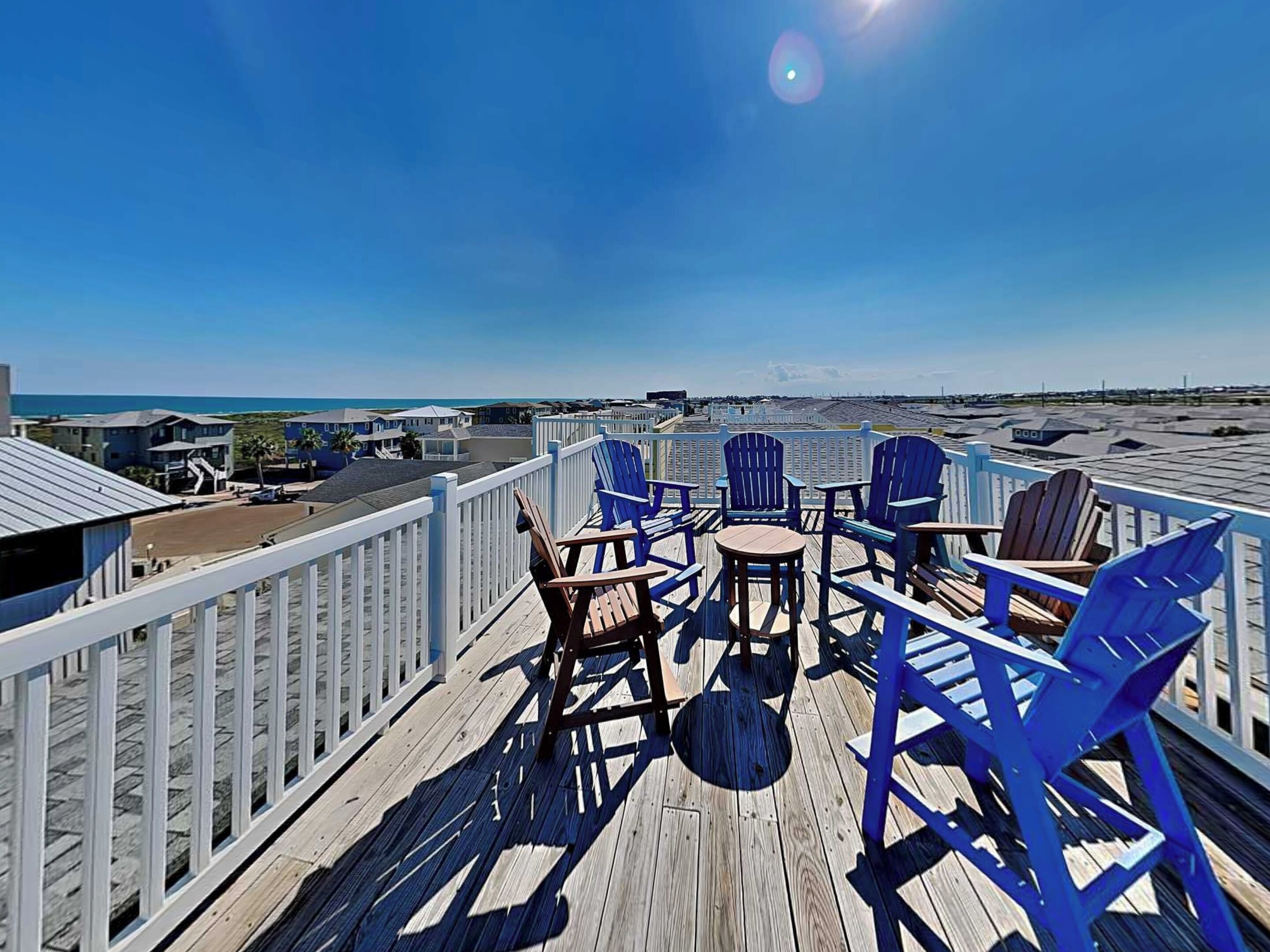 A wooden deck with blue Adirondack chairs and a small round table, overlooking a beach and ocean.