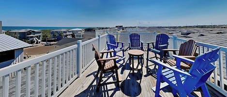 A wooden deck with blue Adirondack chairs and a small round table, overlooking a beach and ocean.