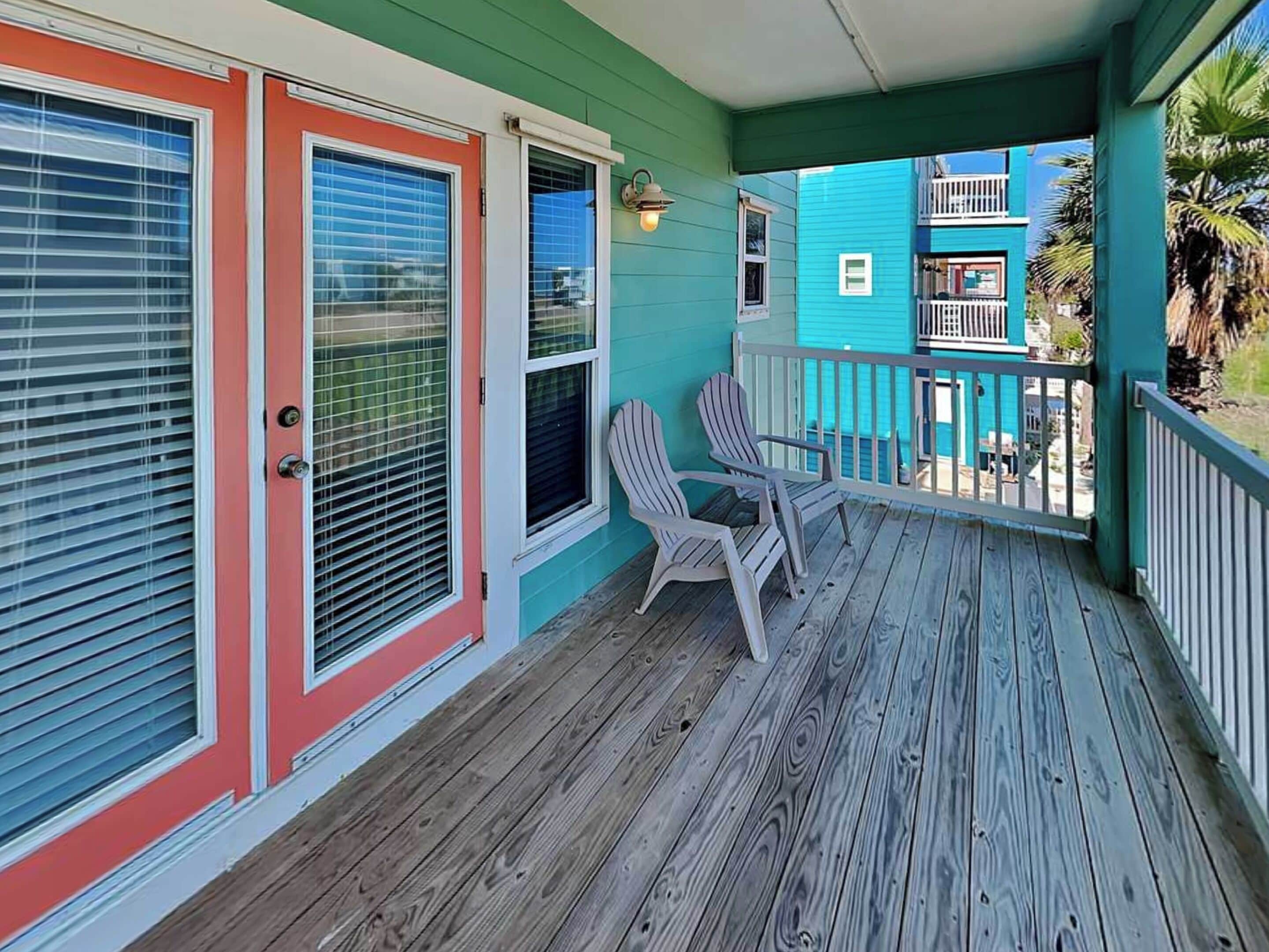 A wooden deck with two Adirondack chairs, a teal-colored building with a balcony, and a red door with a window.