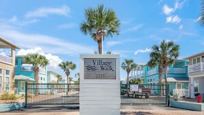 A sign for Village Walk, number 2525, with a gated entrance and palm trees. - The Fire Escape by Silver Sands Vacation Rentals (Port Aransas)