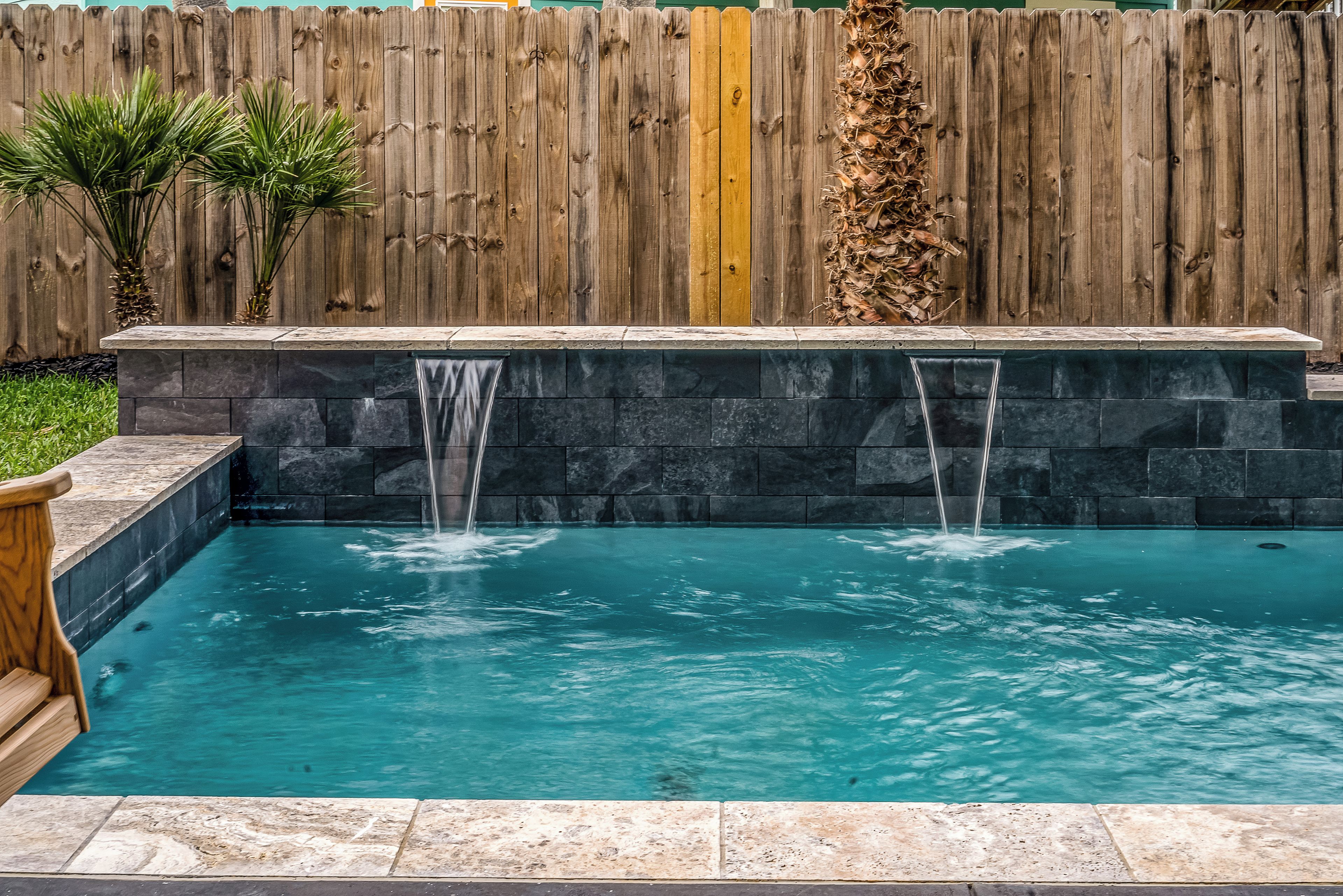 Ein rechteckiger Pool mit zwei Wasserspielen, umgeben von einem Holzzaun und Steinmauern.