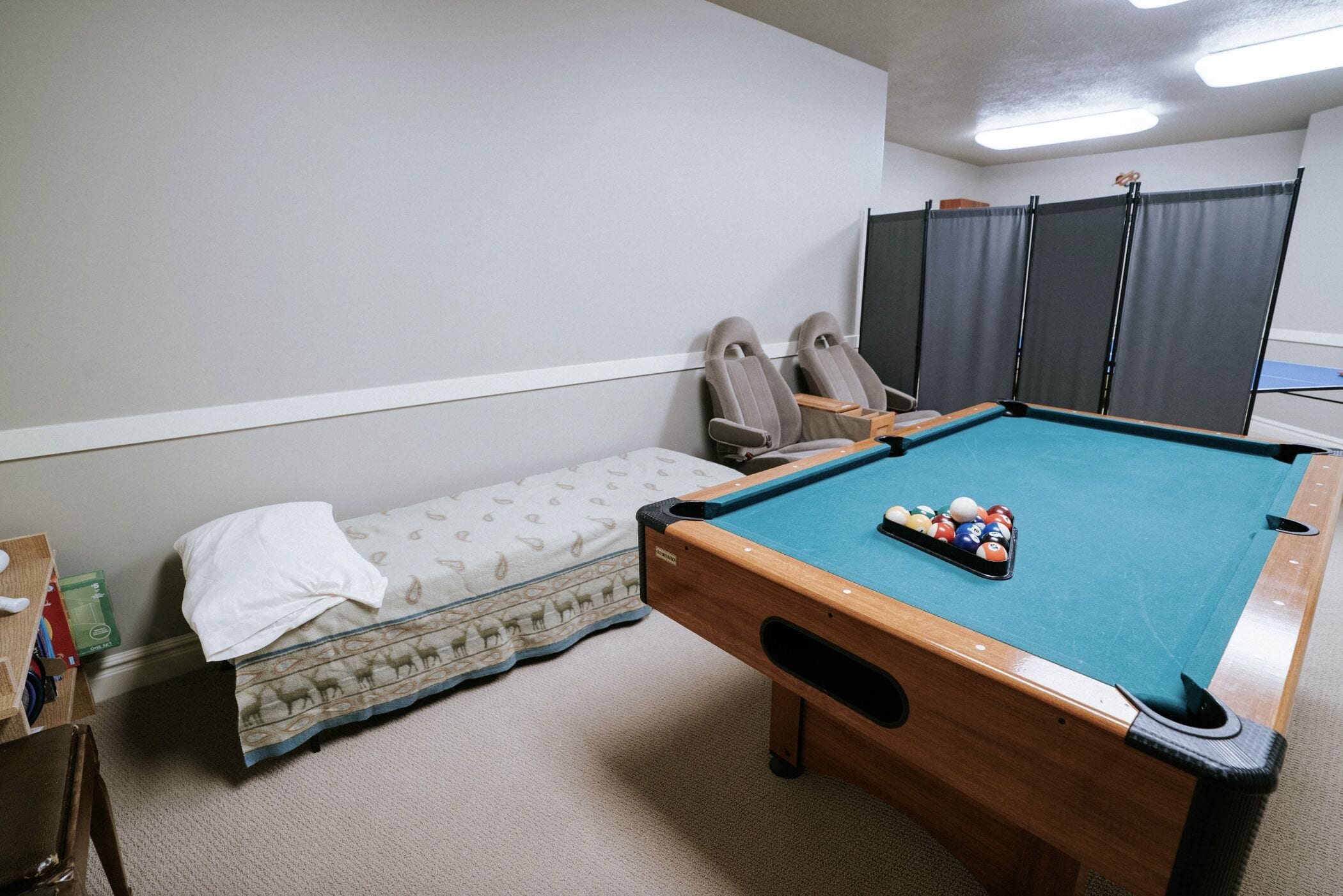 A room with a pool table, two chairs, a bed, and a small table with items on it.