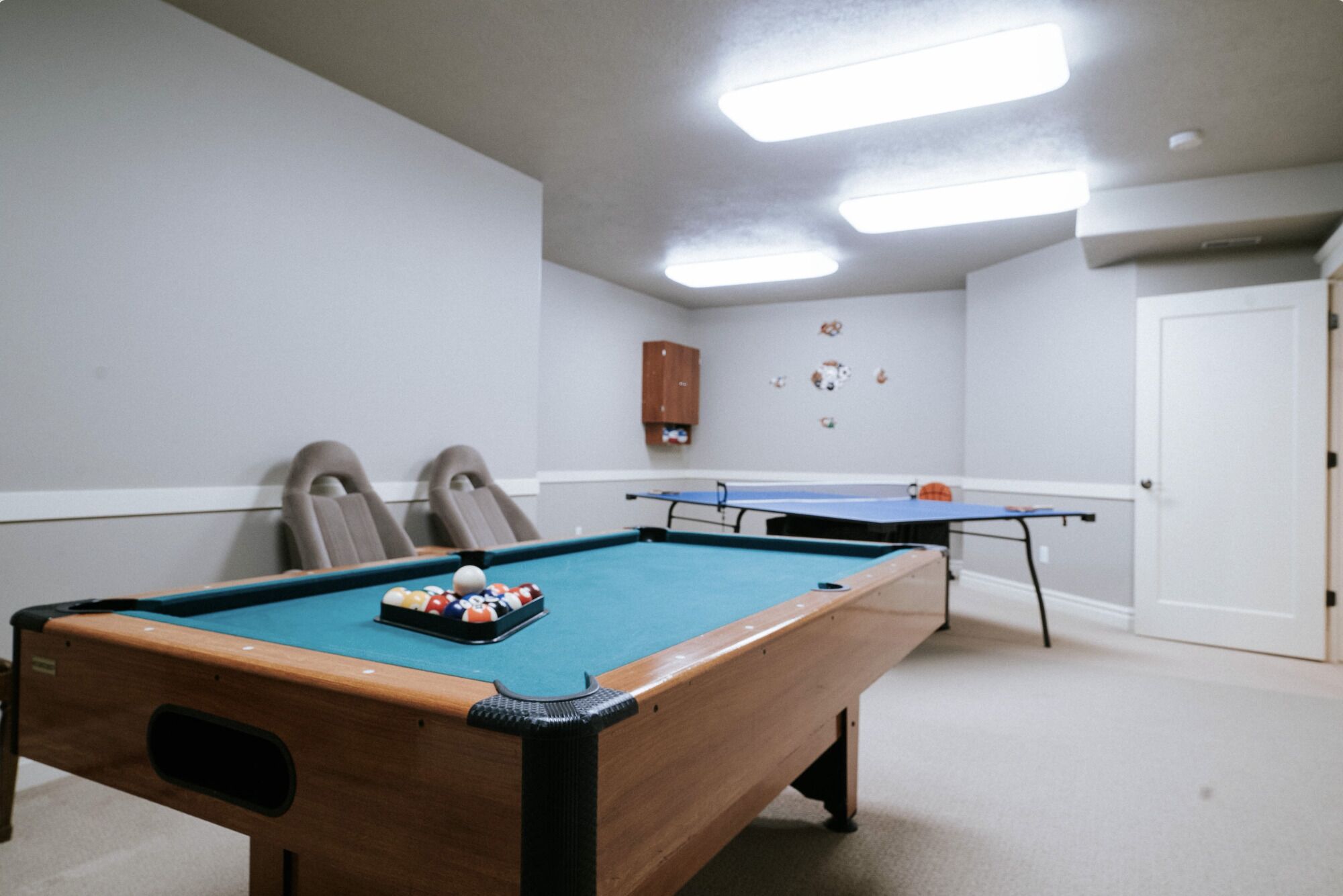 Une table de billard avec des billes au centre d’une pièce, deux chaises et une table de ping-pong à l’arrière-plan.