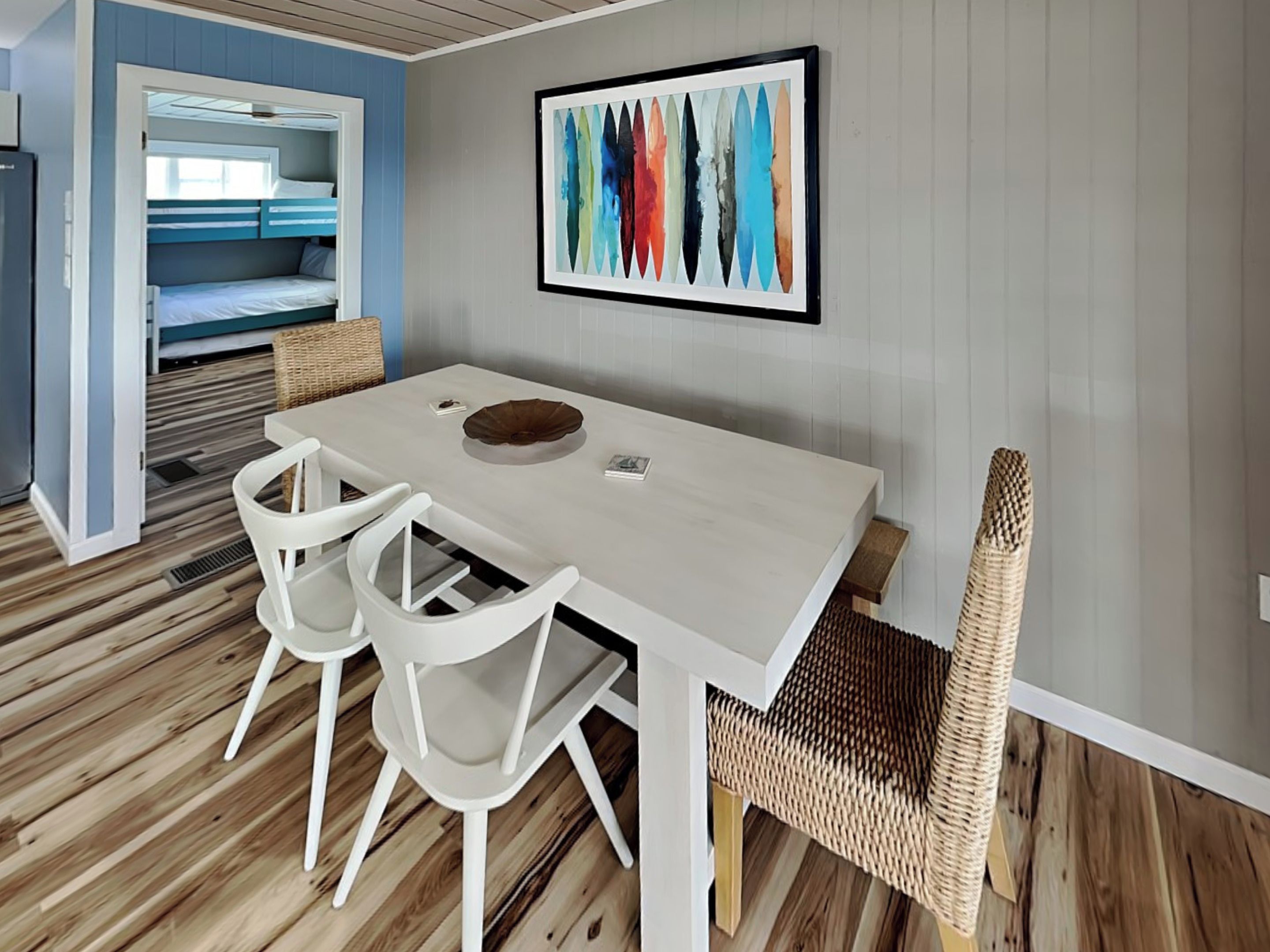 Un'area pranzo moderna con tavolo e sedie bianche, una sedia in rattan e un quadro astratto colorato appeso alla parete.