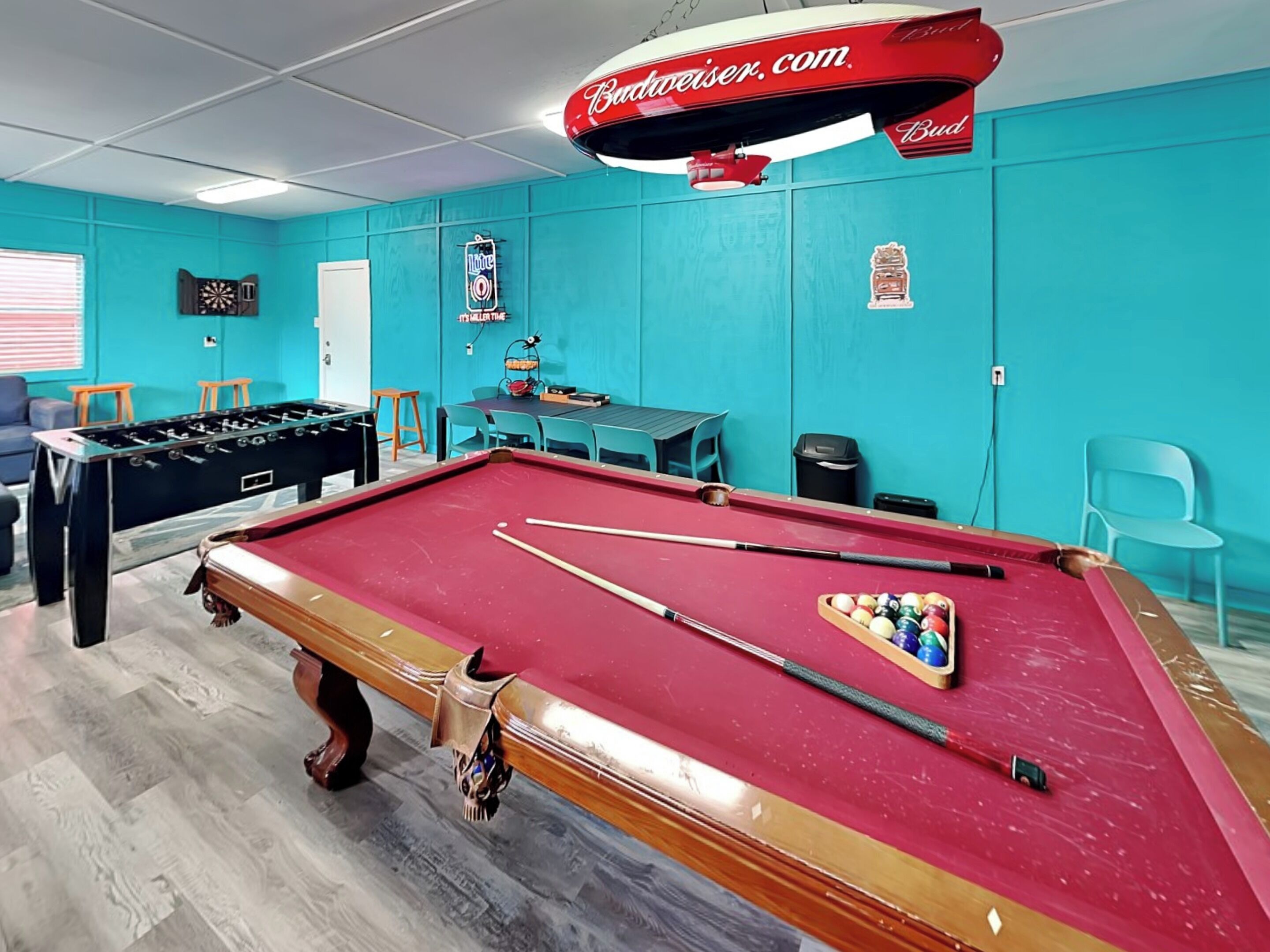 A pool table with pool cues and balls, a foosball table, and a dartboard in a room with teal walls and a ceiling fan.