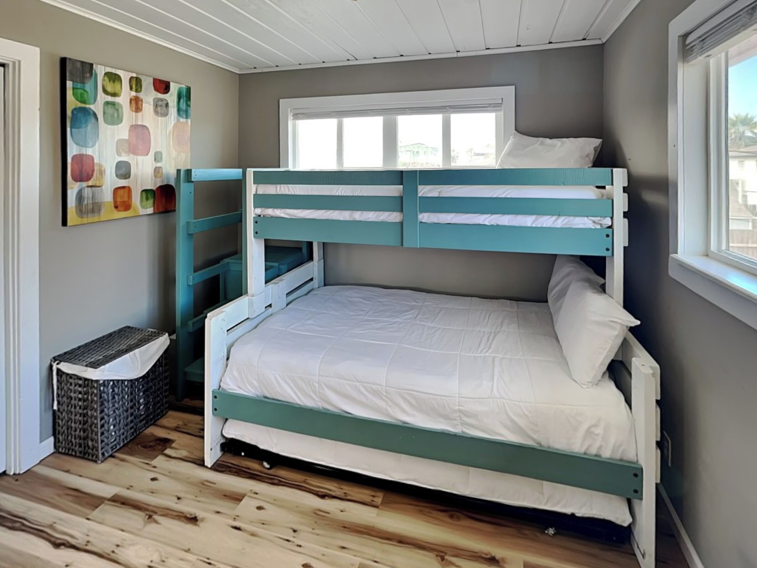 Un lit superposé avec un lit inférieur, une fenêtre avec des stores, un plancher en bois et une peinture abstraite aux couleurs vives accrochée au mur.