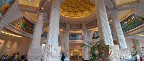 A grand lobby with ornate columns, a high ceiling with decorative patterns, and a large central chandelier.