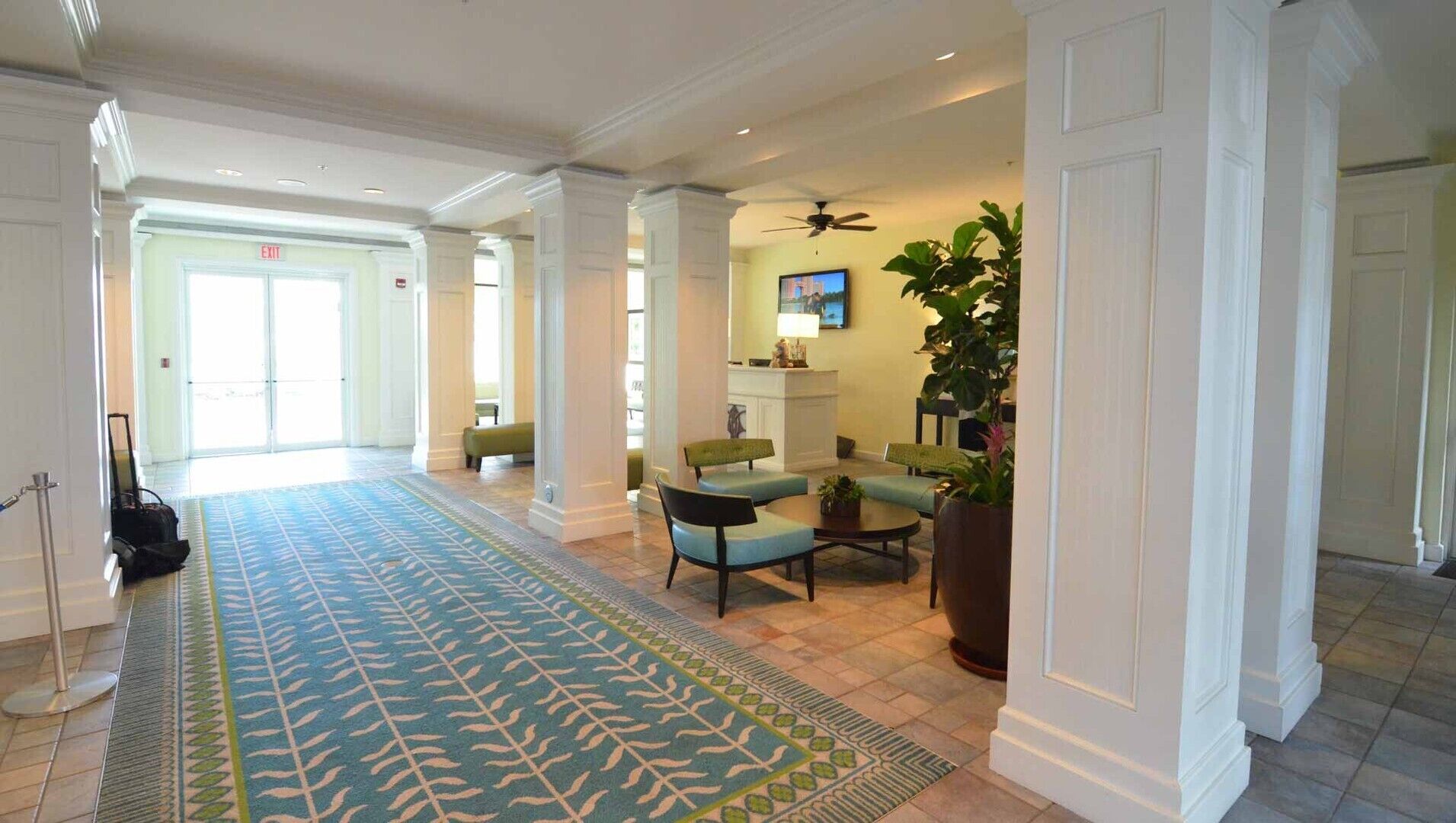 A spacious lobby with a patterned carpet, seating area with chairs and a table, a television mounted on the wall, and a ceiling fan.