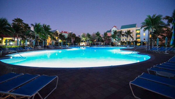 A large outdoor swimming pool surrounded by lounge chairs and palm trees, with hotel buildings in the background.
