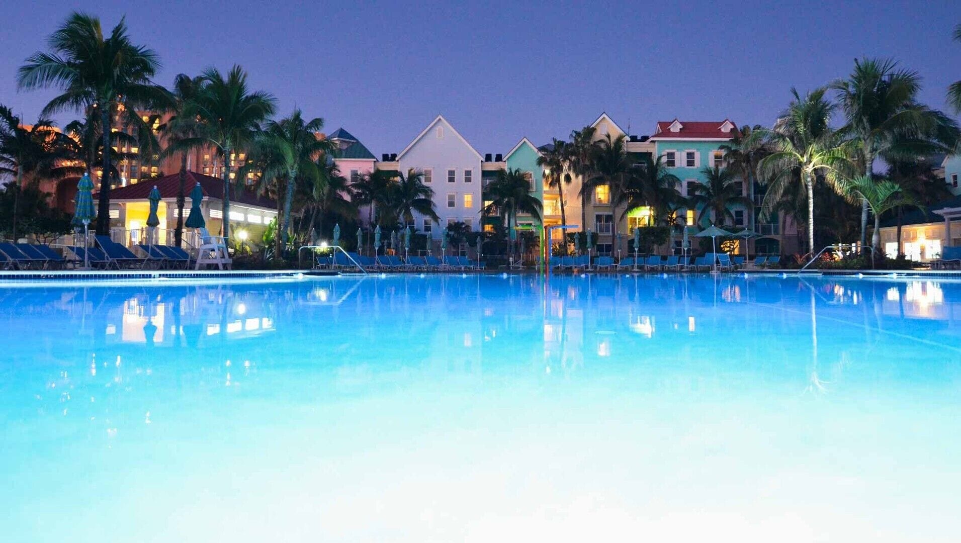A swimming pool with lounge chairs and palm trees, surrounded by colorful buildings.