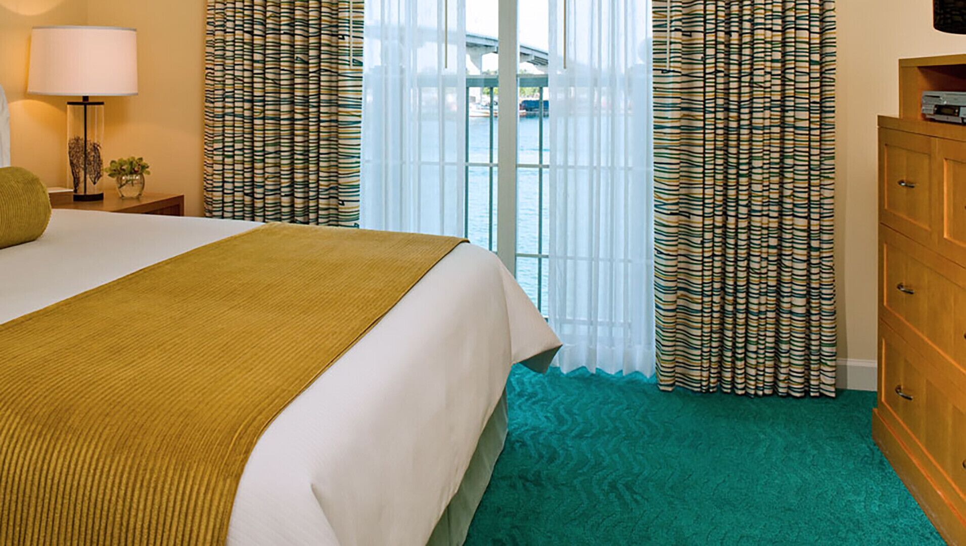 A hotel room with a bed, a wooden dresser, a lamp, and curtains.