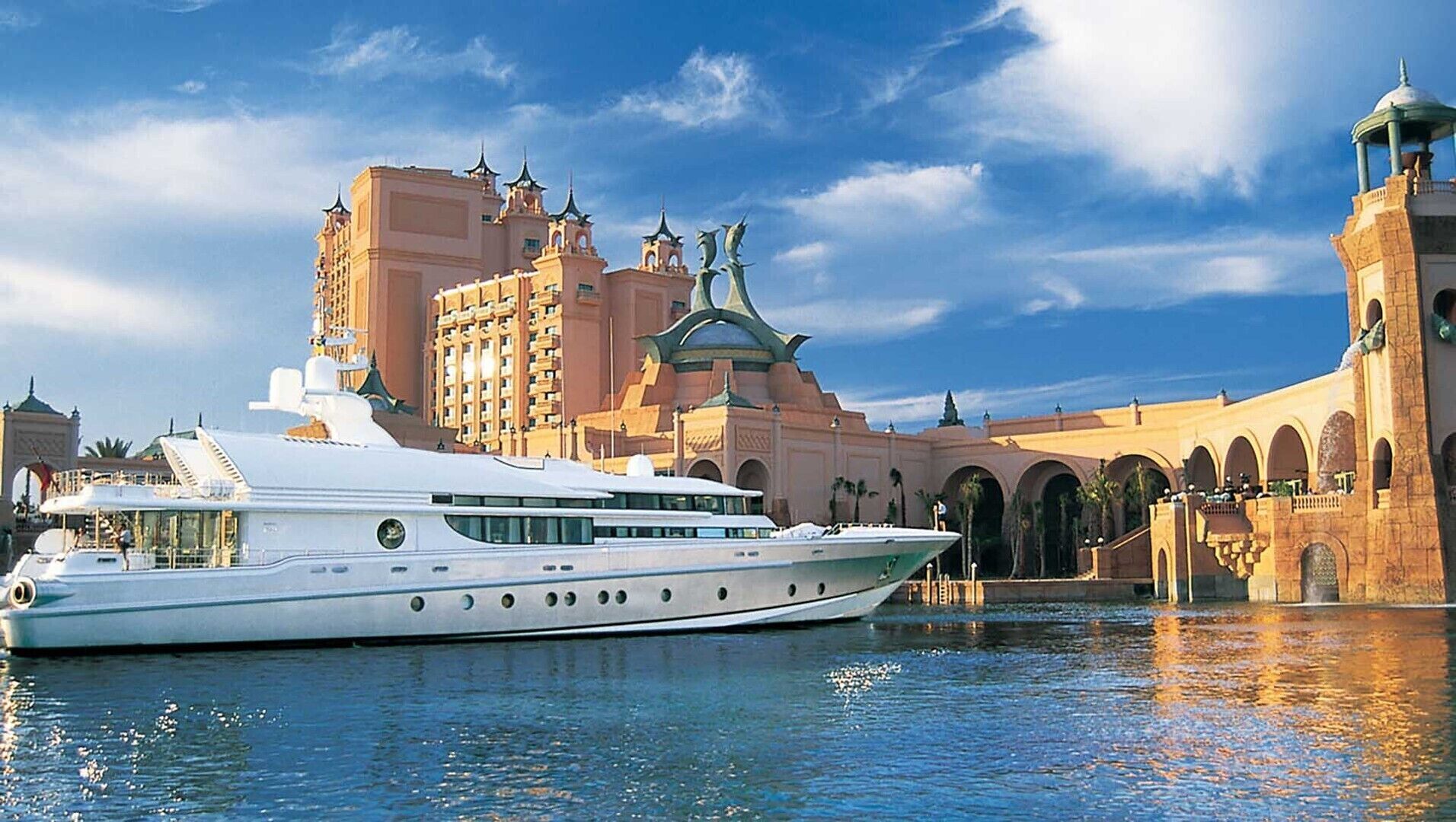 Un grand yacht blanc était amarré devant un imposant bâtiment doté de nombreuses arcades et d’une tour.
